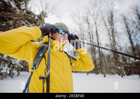 Älterer Mann, der eine Schneebrille aufsetzt und sich auf die Fahrt im verschneiten Wald vorbereitet. Stockfoto