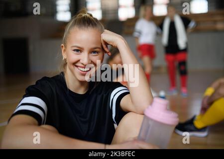 Porträt einer jungen Frau, Sportteamspielerinnen im Fitnessstudio, die nach dem Spiel mit ihren Teamkollegen sitzen und sich ausruhen. Stockfoto