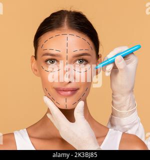 Plastischer Chirurg, der für die kosmetische Operation auf beigem Studiohintergrund Markierungen auf das Gesicht einer jungen Frau zeichnet Stockfoto