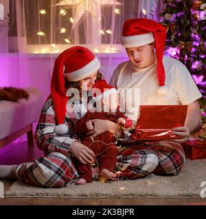 Baby Junge Kleinkind mit seiner Mutter und Vater öffnen Geschenke am Familie Weihnachtsbaum am Silvesterabend. Glückliches Kind in den Händen einer Frau in einem santa ha Stockfoto