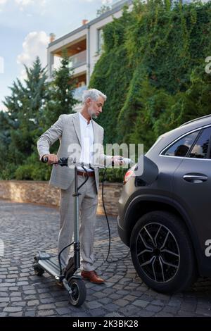 Nahaufnahme eines Geschäftsmannes auf dem Weg zur Arbeit, der am Elektroscooter steht und sein Elektroauto lädt. Konzept des ökologischen Pendelns und der Umweltfreundlichkeit Stockfoto