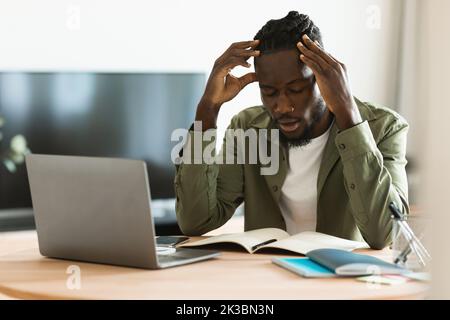 Trauriger schwarzer Freiberufler, der am Arbeitsplatz einen Laptop ansieht, an Kopfschmerzen leidet oder Probleme hat Stockfoto