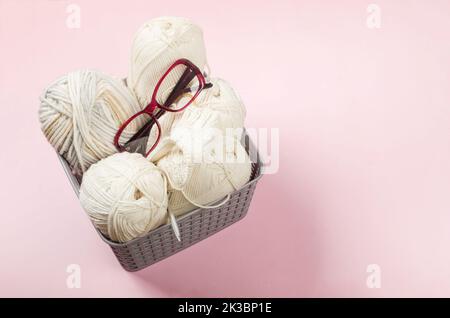 Hellbeige Strickwolle, Stricknadeln und Sehnen im Korb auf pastellrosa Hintergrund. Weibliche Hobby Stricken. Weiche Wollgarne zum Stricken Stockfoto