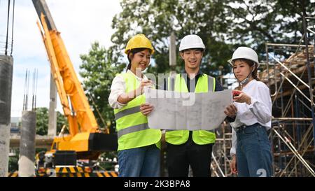 Die Manager des Bauingenieurs im Schutzhelm besprechen die Plandetails, während sie auf der Baustelle mit einem Kran im Hintergrund stehen Stockfoto