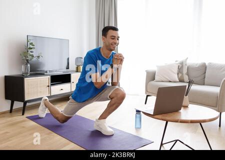 Mann, der zu Hause trainiert. Fröhlicher junger arabischer Kerl, der Übungen für Beine im Wohnzimmer macht und auf einen Laptop schaut Stockfoto