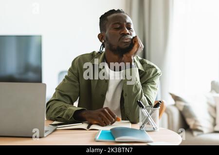 Nachdenklicher afroamerikanischer Mann, der am Laptop arbeitet und über den Job nachdenkt, am Arbeitsplatz im Heimbüro sitzt Stockfoto