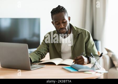 Fokussierter afroamerikanischer Mann, der am Schreibtisch sitzt und am Laptop arbeitet und sich im Notebook Notizen macht, um zu Hause online zu studieren Stockfoto