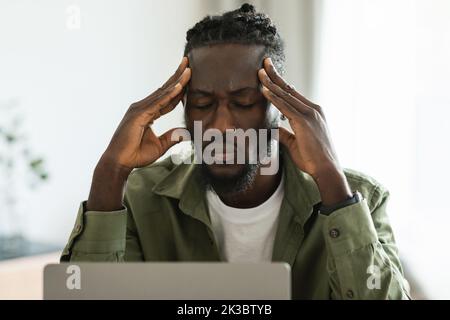 Unglücklicher schwarzer freiberuflicher Mann, der den Kopf berührt, Kopfschmerzen hat oder Probleme mit der Arbeit hat, am Schreibtisch sitzt Stockfoto