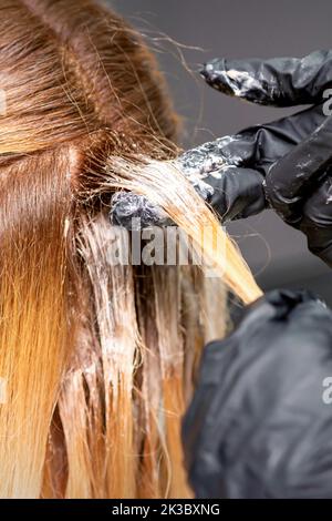 Die Hände des Friseurs tragen den Farbstoff mit Handschuhen auf das rote Haar einer jungen rothaarigen Frau in einem Friseursalon aus nächster Nähe auf Stockfoto