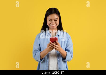 Lächelnd hübsche junge chinesische Studentin beim gelegentlichen Tippen auf dem Smartphone, Surfen im Internet, Video ansehen Stockfoto