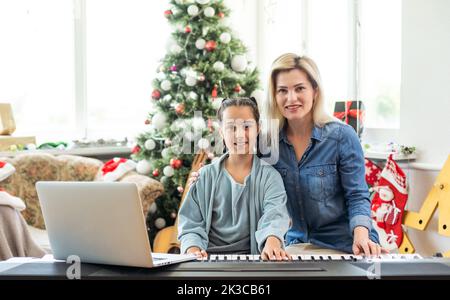 Porträt eines kleinen Mädchens, das Klavier spielt und mit Weihnachtsornamenten verziert ist. Glückliche Mutter lächelt auf dem Hintergrund. Winter- und Neujahrsferien Stockfoto