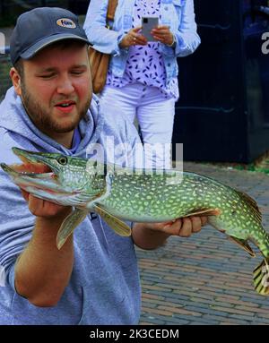 Elburg, Niederlande, September 23 2022, stolzer Fischer, der seinen gefangenen Hecht zeigt. Im Hintergrund eine Frau, die mit ihrem Smartphone fotografiert Stockfoto