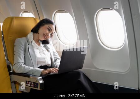 Fröhliche asiatische Geschäftsfrau telefonieren und mit Laptop-Computer während des Fluges. Urbane Lebensart Stockfoto