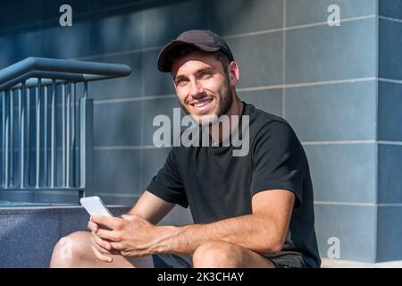 Fröhlicher junger Mann in schwarzem T-Shirt und Hut, der durch sein Handy surft und mit einem Lächeln auf die Kamera blickt, während er auf einer Treppe vor dem modernen Gebäude sitzt Stockfoto