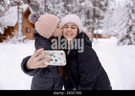 Glückliche, liebevolle Schwestern küssen und machen Selfie im Schnee