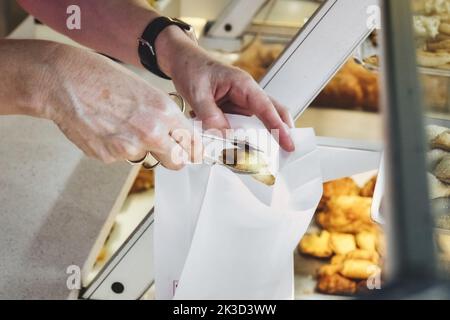 Nahaufnahme der Hände einer Person, die eine Teigzange hält, die einen süßen Kuchen aufnimmt und ihn in einen weißen Papierbeutel legt Stockfoto