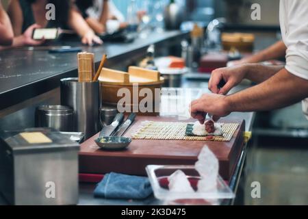 Nahaufnahme der Hände eines Küchenchefs, der an einer Sushi-Bar eine Sushi-Rolle mit Reis und Algen rollt, mit Leuten im Hintergrund an der Theke Stockfoto