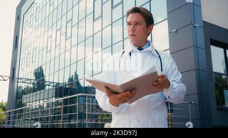 Doktor in weißem Mantel, der Ordner hält und in der Nähe des Gebäudes des Krankenhauses wegschaut, Stockbild Stockfoto