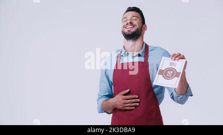 Aufgeregter Kellner lachte und berührte den Bauch, während er das Menü isoliert auf einem grauen Bild hielt Stockfoto