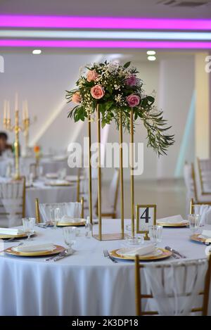 Ein Tisch mit Geschirr und Blumenarrangements auf Ständen in einem Hochzeitsbankettssaal Stockfoto