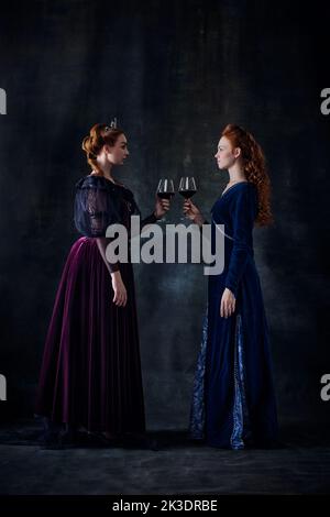 Porträt von zwei schönen Frauen im Bild von Königinnen stehen von Angesicht zu Angesicht mit einem Glas Wein isoliert auf dunklem Hintergrund Stockfoto