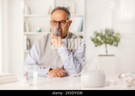 Reifer Mann sitzt auf einem Tisch und benutzt zu Hause eine Inhalationsmaschine Stockfoto