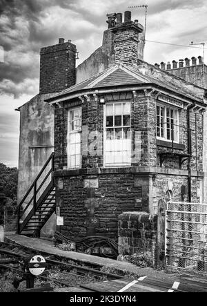 An einer Kreuzung zwischen Straße und Schiene steht eine alte Backsteinsignalbox. Eine Treppe führt nach oben ins Innere und Schienen sind im Vordergrund. Stockfoto