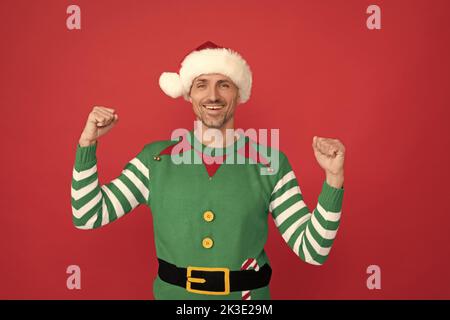 Glücklicher erfolgreicher Mann im Elf Kostüm. weihnachtsmann im weihnachtsmann Hut auf rotem Hintergrund. Stockfoto