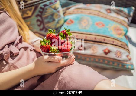 Portrait einer wunderschönen Frau in einem wunderschönen Kleid, die auf einer Terrasse sitzt und Erdbeere isst Stockfoto