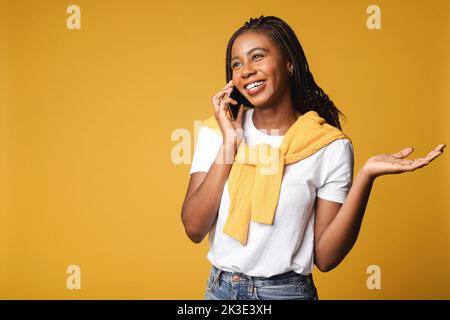 Zufriedene, sorglose afroamerikanische Frau mit Dreadlocks, spricht per Smartphone, hat ein angenehmes Telefongespräch mit einem Freund, genießt das Sprechen mit jemandem, isoliert auf Gelb Stockfoto