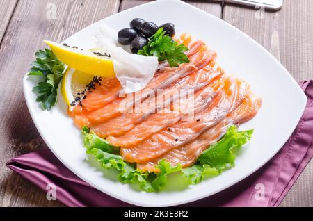 Frischer roter Fisch, der auf einem weißen Teller auf dunklem Holzgrund in Streifen mit Zitrone und Kräutern geschnitten wurde Stockfoto