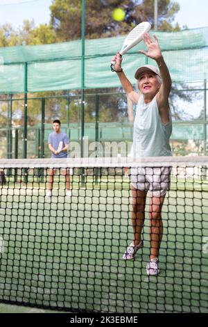 Ältere Frau, die beim Paddleball-Spiel auf dem Platz im Freien den Ball serviert Stockfoto