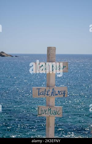 Blick auf hölzerne Schilder für Take Away, Cold Brew, Live-Musik und das Meer im Hintergrund am Strand von Mylopotas in iOS Griechenland Stockfoto