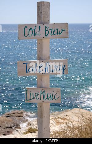 Blick auf hölzerne Schilder für Take Away, Cold Brew, Live-Musik und das Meer im Hintergrund am Strand von Mylopotas in iOS Griechenland Stockfoto