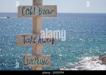 Blick auf hölzerne Schilder für Take Away, Cold Brew, Live-Musik und das Meer im Hintergrund am Strand von Mylopotas in iOS Griechenland Stockfoto