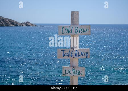 Blick auf hölzerne Schilder für Take Away, Cold Brew, Live-Musik und das Meer im Hintergrund am Strand von Mylopotas in iOS Griechenland Stockfoto