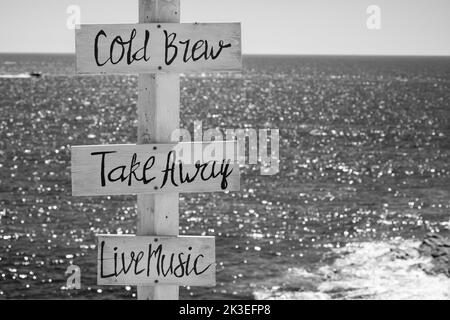 Blick auf Holzschilder für Take Away, Cold Brew, Live-Musik und das Meer im Hintergrund am Strand von Mylopotas in iOS Greece in Schwarz und Weiß Stockfoto