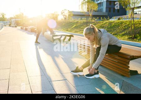 Junge blonde Sportlerin in Sportbekleidung sitzt auf der Bank in städtischer Umgebung und beugt sich über Sneaker, während sie Schnürsenkel bindet Stockfoto