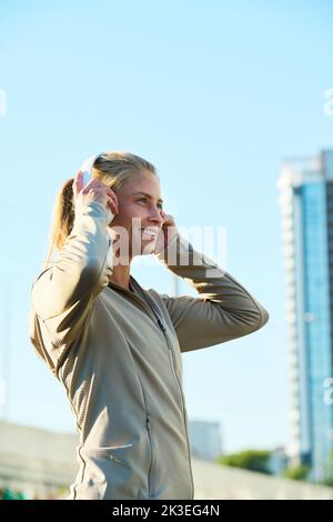 Seitenansicht einer jungen, lächelnden Sportlerin, die in Kopfhörern Musik hört, während sie in der Stadt vor der Kamera steht Stockfoto