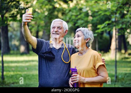 Altes sportliches Paar starrt auf Smartphone-Kamera Selfie-Foto machen Genießen Sie die moderne Nutzung von Wireless-Technologie, während Sie in Sportkleidung im Sommerpark spazieren. Carefre Stockfoto