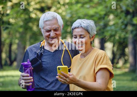 Attraktive ältere, grauhaarige Ehefrau und Ehemann verwenden ein modernes Smartphone, um den Kalorienzähler zu überprüfen oder nach sportlichem Sport im Sommerpark zu verfolgen. Tec Stockfoto