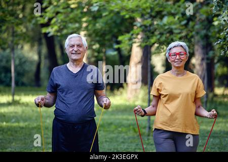 Ältere Ehegatten tragen Sportkleidung und machen morgens im Sommerpark Übungen mit Gummibändern. Gesunder Lebensstil, aktiver Ruhestand Stockfoto
