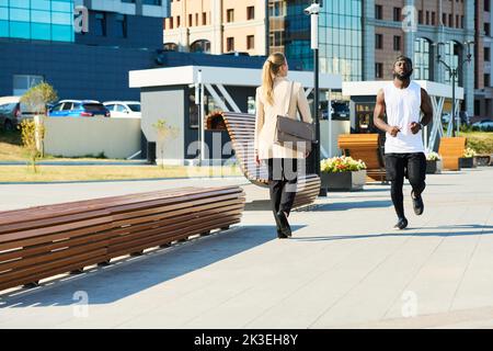 Der junge Sportler joggt auf der Straße im Park, während die blonde Geschäftsfrau in formalwear am Morgen an ihm vorbeikommt Stockfoto