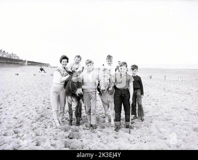 1962, historisch, posiert für ein Foto, eine Mutter, die mit mehreren jungen Jungen steht, die Eselreiten auf einem Sandstrand, geführt von teo-Jugendlichen in Bridlington, Hull, England, Großbritannien. Stockfoto