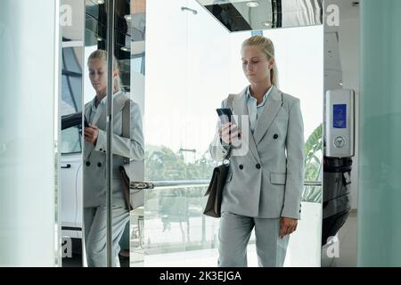Junge, moderne Geschäftsfrau in grauer formalwear, die im Mobiltelefon SMS schreibt, während sie aus dem Aufzug im zweiten Stock des Bürozentrums geht Stockfoto