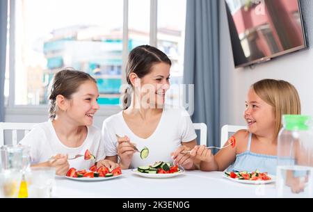 Zwei lustige Mädchen mit Mama essen Gemüsesalat am Esstisch Stockfoto