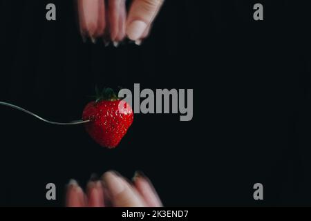 Erdbeere steckt in einer Gabel auf schwarzem Hintergrund. Die Finger der Frau sind verschwommen. Fotostudio. Zugeschnitten auf Werbekampagnen. Stockfoto