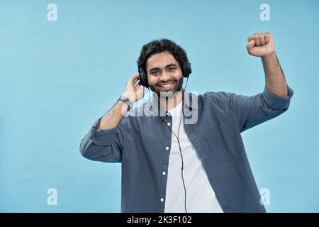 Glücklicher arabischer Mann, der Kopfhörer trägt und Musik hört, die isoliert auf Blau tanzt. Stockfoto