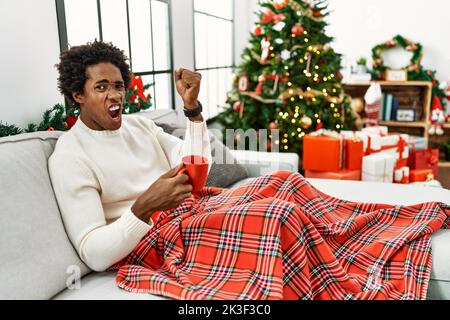 Junger afroamerikanischer Mann, der auf dem Sofa sitzt und am Weihnachtsbaum Kaffee trinkt, wütend und wütend die Faust raffend, frustriert und wütend, während er mit ihm schrie Stockfoto