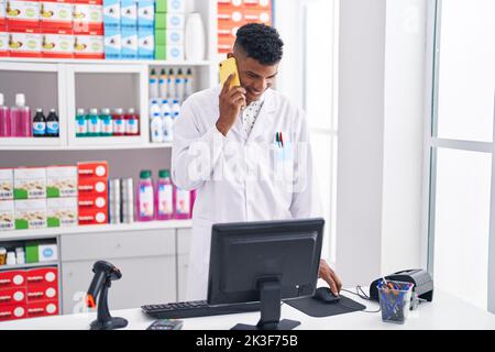 Junger latein-Apotheker im Gespräch auf dem Smartphone mit Computer in der Apotheke Stockfoto